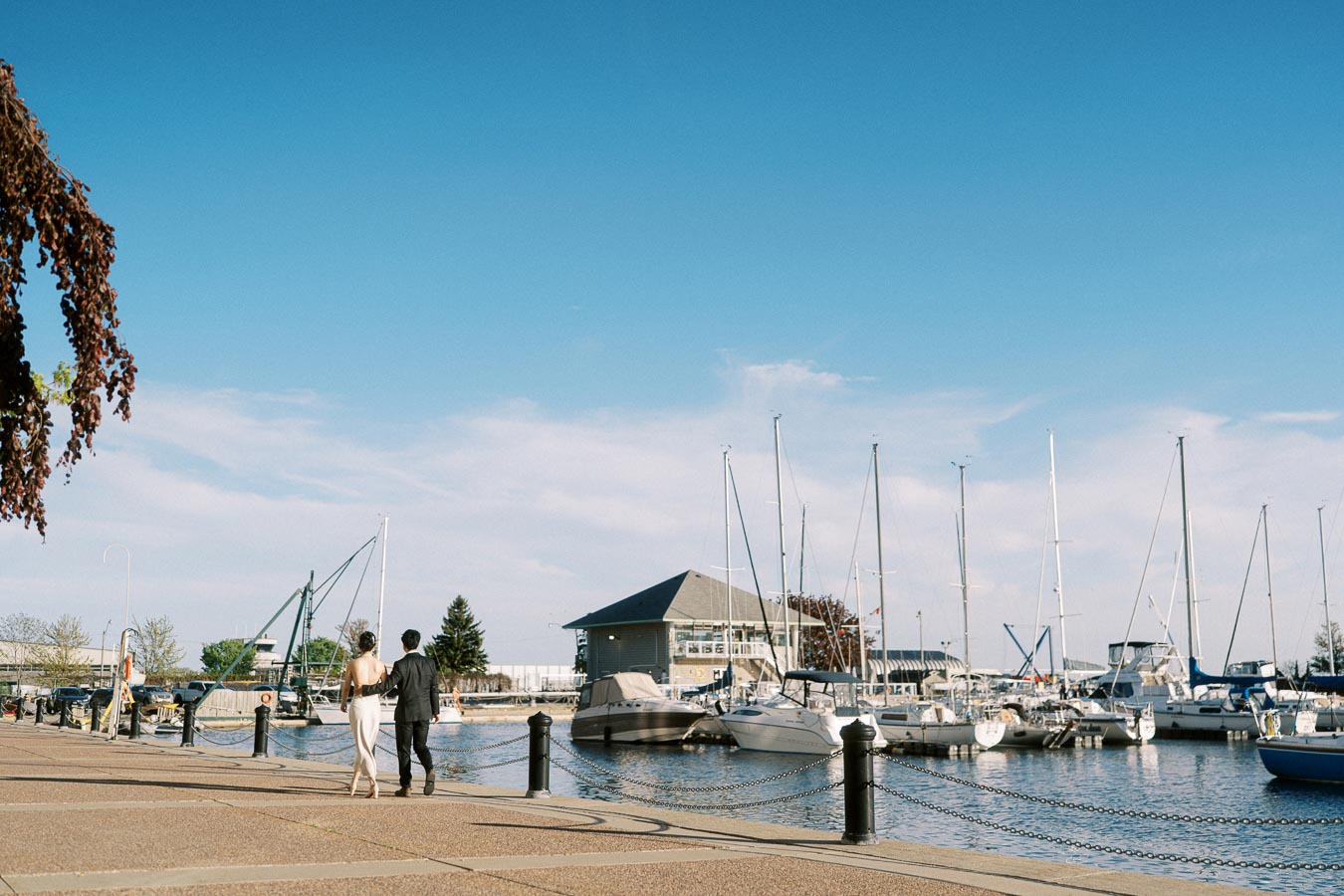 Engaged couple walking hand in hand along the marina boardwalk at Trillium Park in Toronto, with sailboats and a clear spring sky in the background.