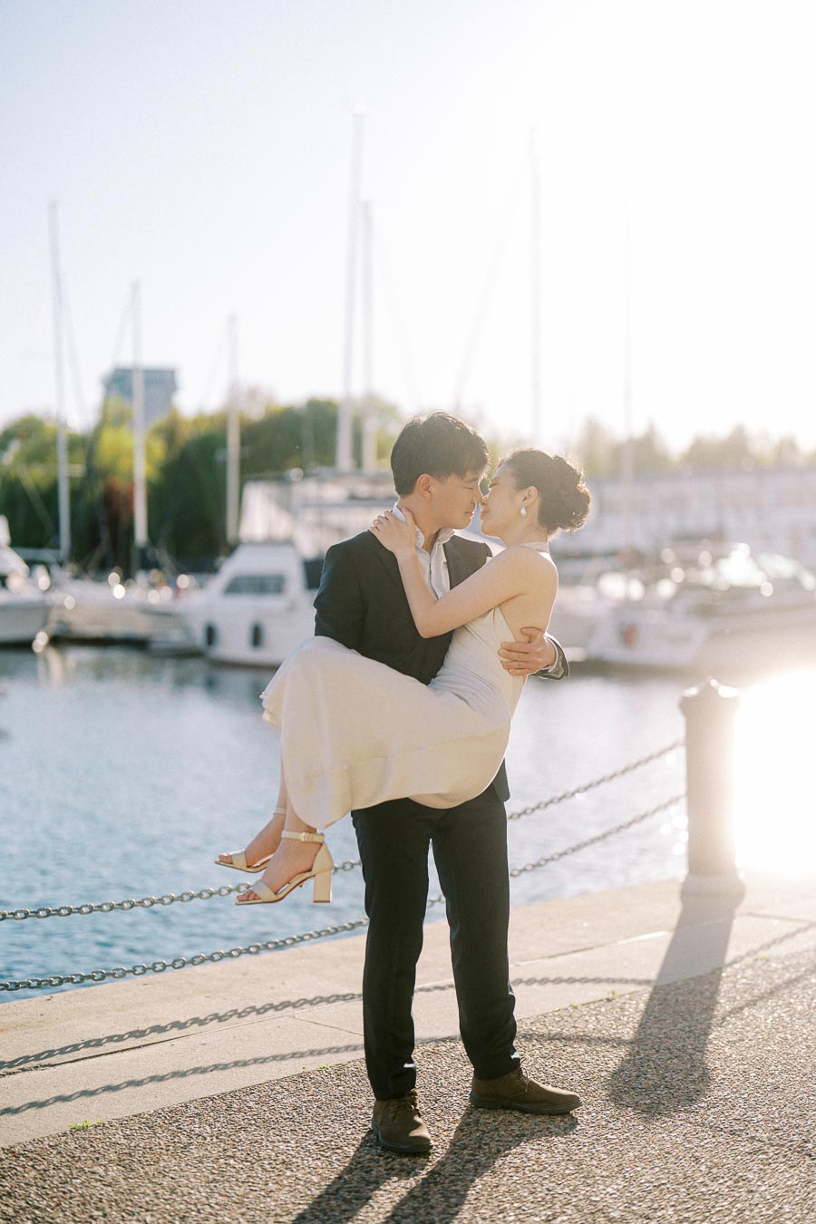 Engaged couple at Trillium Park, Toronto, with the groom-to-be lifting his bride-to-be in front of a sunlit marina and sailboats along the waterfront.