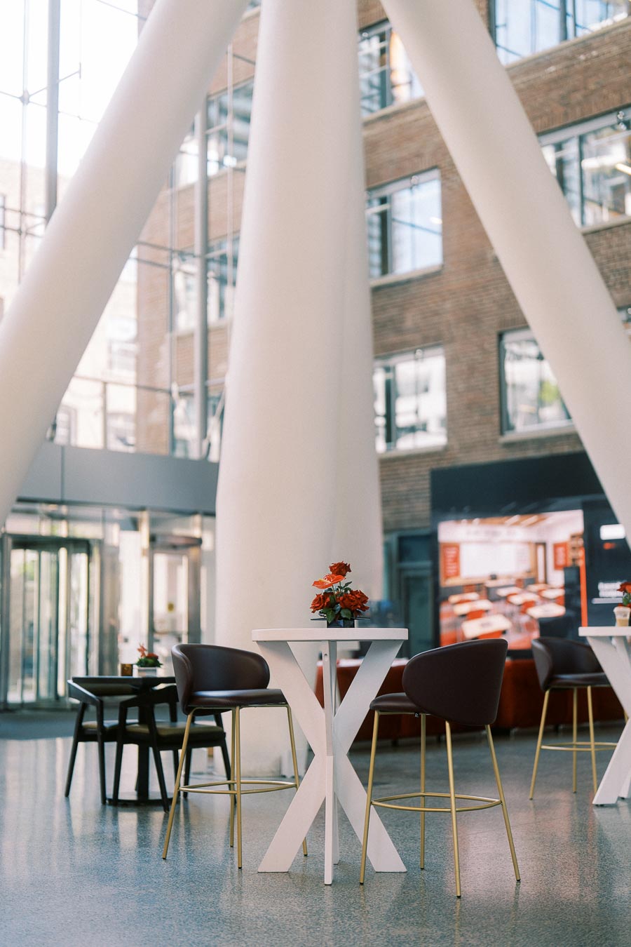 High-top tables with red floral arrangements and modern chairs in the bright, spacious Atrium at Ricarda’s Toronto, featuring architectural columns and brick walls.