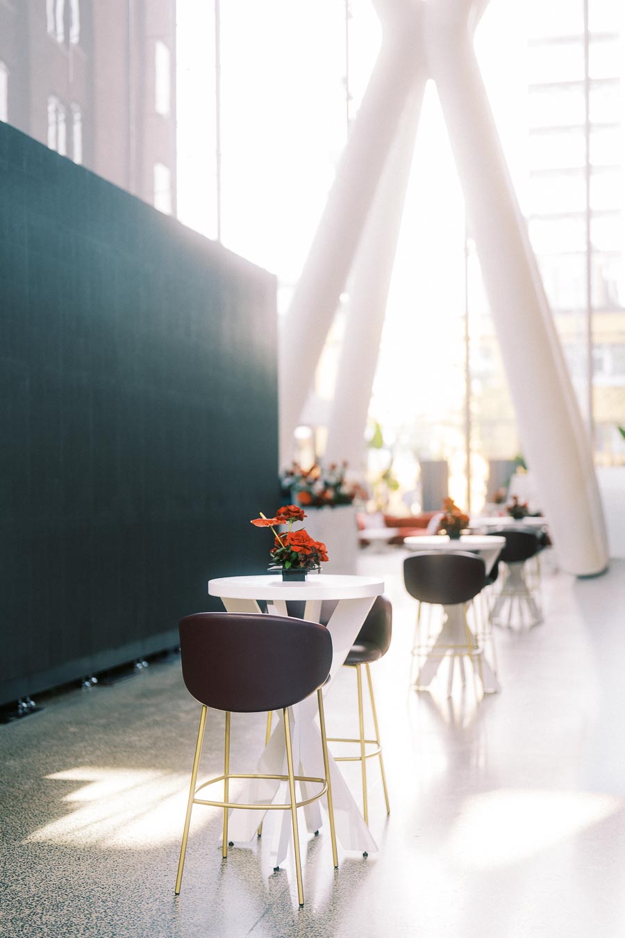 Modern white cocktail tables with dark chairs and red floral centrepieces in the bright atrium of Ricarda’s, Toronto, set for a school launch event.
