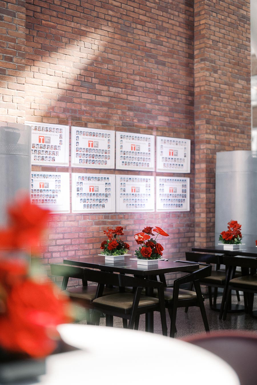Tables with red floral centrepieces set against exposed brick walls and Toronto Prep School class photo displays at Ricarda’s Atrium in Toronto.