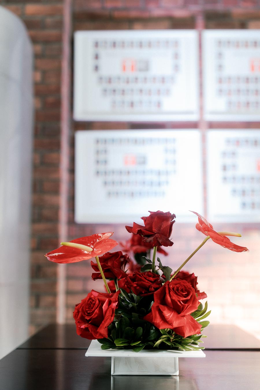Red roses and anthuriums arranged in a modern centerpiece on a table at Ricarda’s Restaurant in Toronto, with a blurred brick wall and signage in the background.