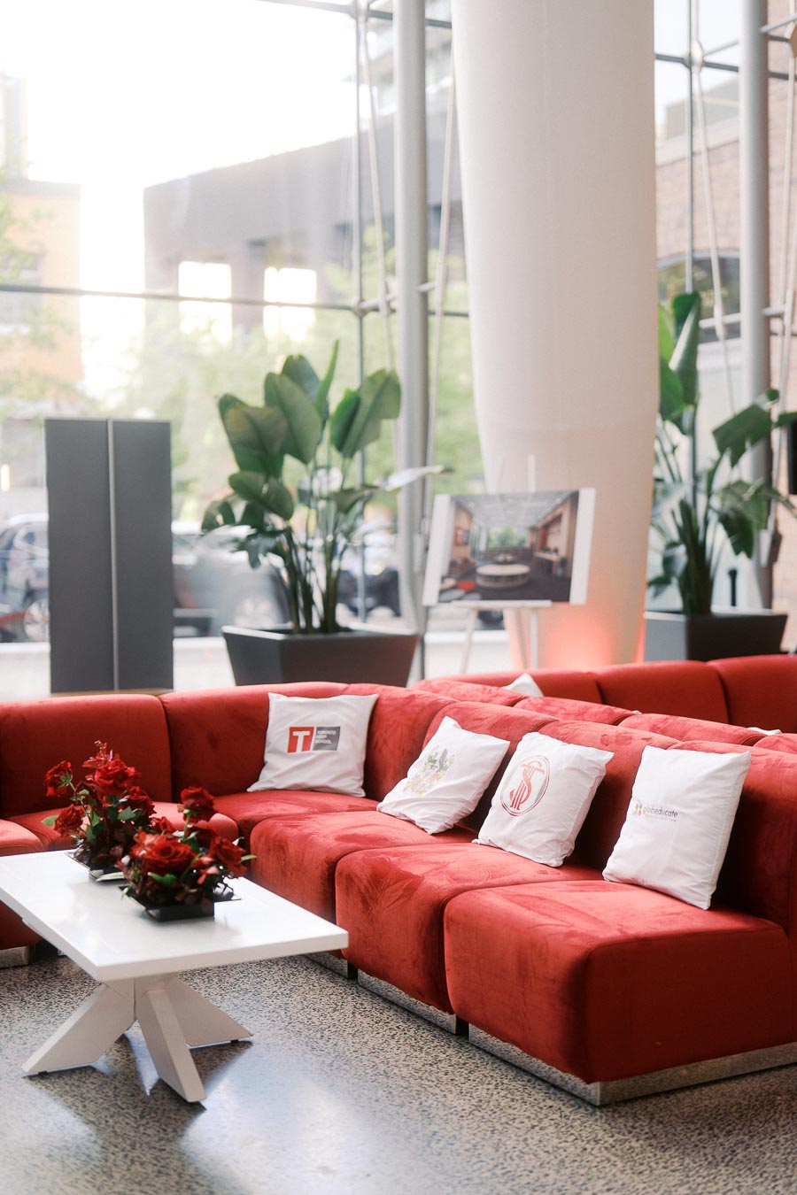 Red sectional sofa with Toronto Prep School and Globeducate branded pillows, white coffee table with red floral arrangements, and modern atrium windows at Ricarda’s, Toronto.