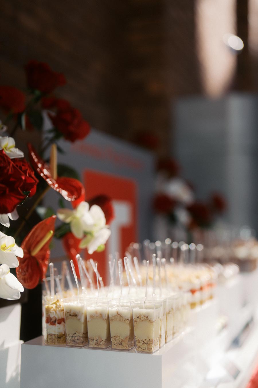 Rows of individual dessert cups with spoons on a white stand, decorated with red and white flowers, at Ricarda's in Toronto during a school event.