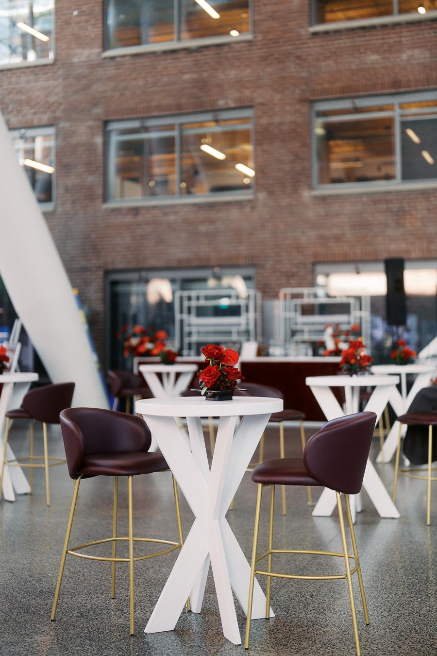 White cocktail tables with red rose floral centrepieces and burgundy chairs arranged in the modern atrium of Ricarda’s, Toronto, during the Toronto Prep Forever Launch Party.