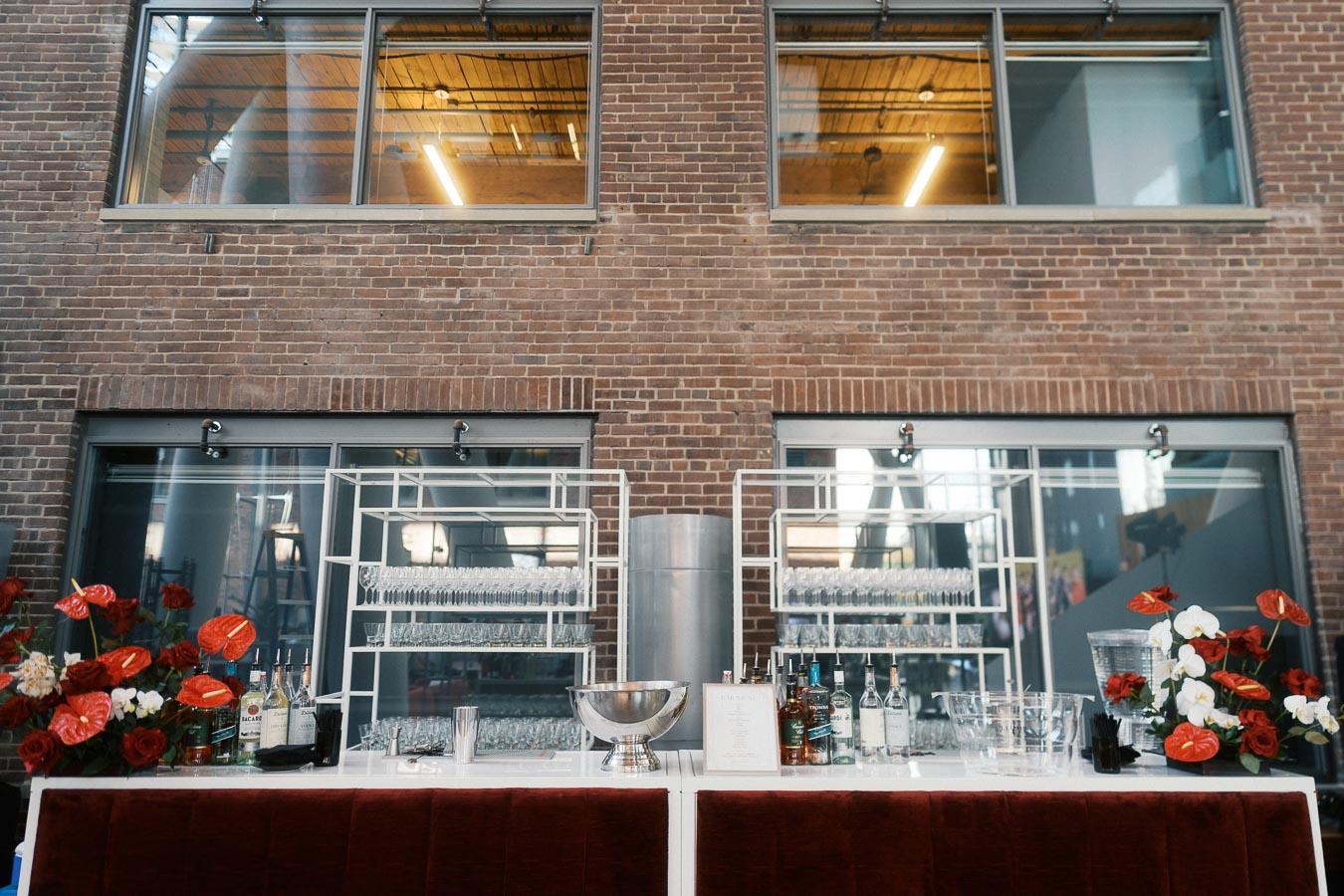 A modern bar setup with red and white flowers, bottles, glassware, and a brick wall backdrop at Ricarda’s in Toronto during the Toronto Prep Forever Launch Party.