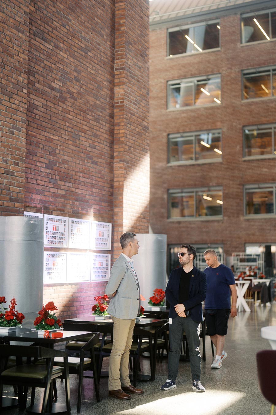 Two men talking near a brick wall and decorated tables at The Atrium at Ricarda’s in Toronto during the Toronto Prep Forever Launch Party. A third guest walks by in the background.