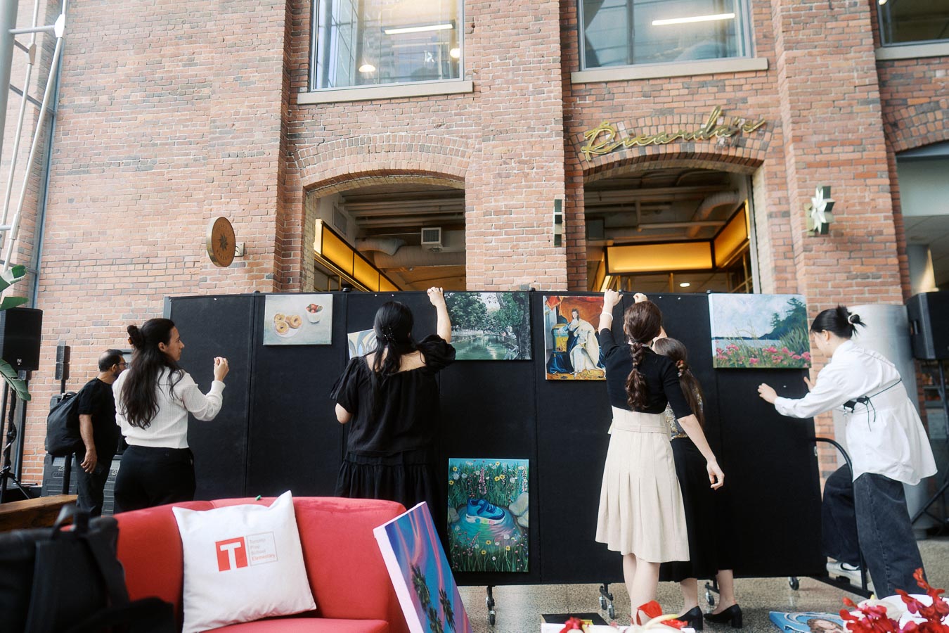 Five people arranging paintings on a black display wall inside Ricarda’s, Toronto, with exposed brick walls and event signage visible above.