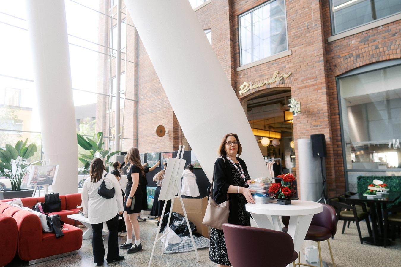 A woman stands by a table with event materials as guests view school artwork in the modern atrium of Ricarda’s Restaurant during the Toronto Prep School launch event.