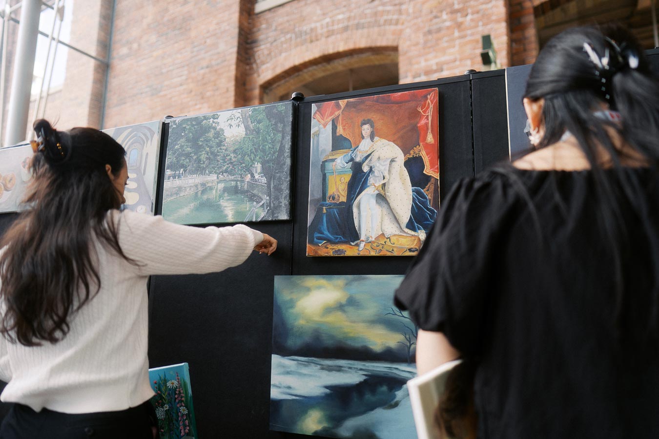 Two people stand in front of a display of student artwork at Ricarda’s in Toronto, examining a variety of paintings during a school event.