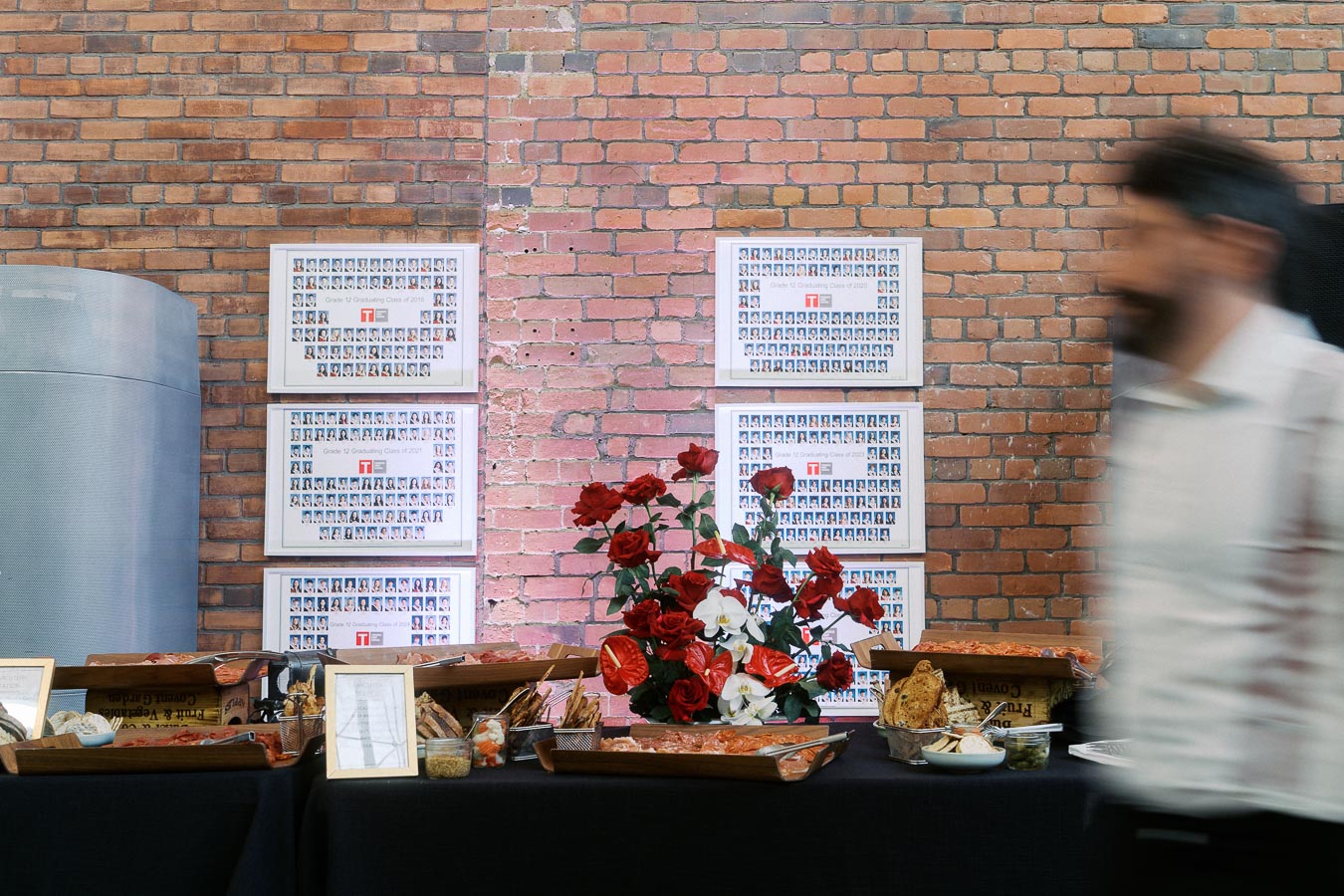 Charcuterie table with floral centrepiece, framed Toronto Prep School class photos on exposed brick wall, Ricarda’s Toronto, person walking by.