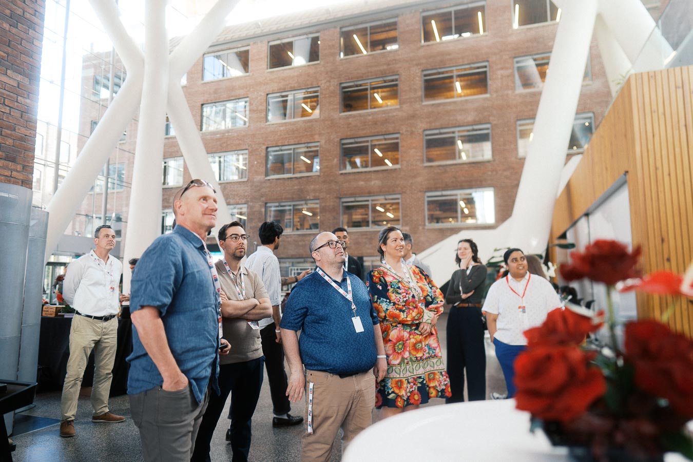 A diverse group of adults stands together in The Atrium at Ricarda’s, Toronto, attentively watching a presentation during the Toronto Prep School Forever Launch Party. Visible are modern architectural elements and a brick building backdrop.