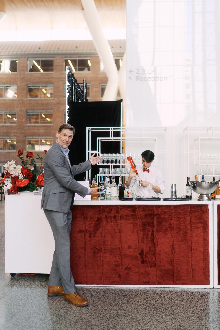 A guest in a grey suit stands at a modern bar with red velvet paneling, gesturing toward a bartender in a white shirt and red bow tie preparing a cocktail at Ricarda’s in Toronto.