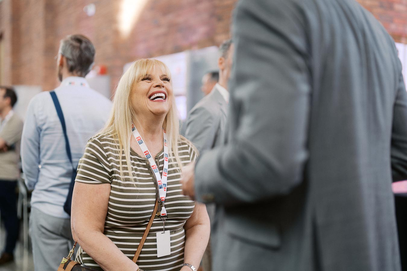 A woman with blonde hair, striped shirt, and TPS lanyard laughs while networking at Toronto Prep Forever Launch Party at Ricarda’s Atrium in Toronto.
