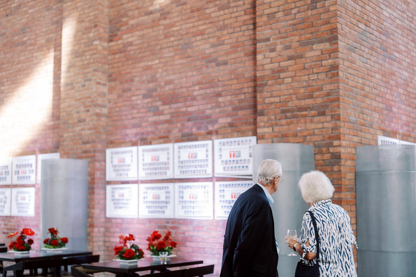 Two older adults stand near alumni photo displays on a brick wall at Ricarda’s in Toronto, with tables decorated with red floral centrepieces in the foreground.