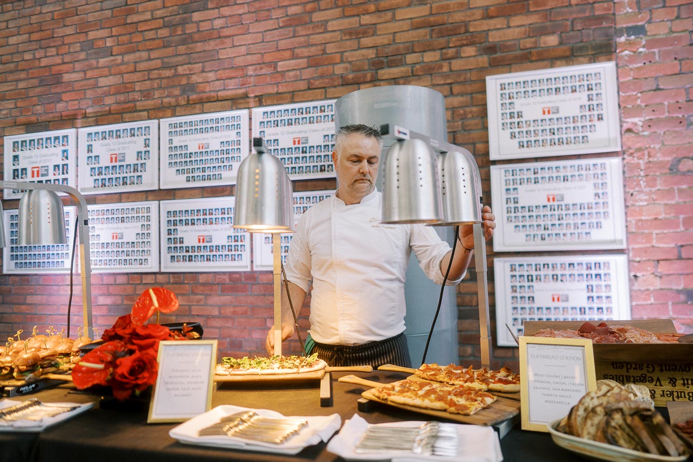 Chef preparing flatbreads under heat lamps at Ricarda’s in Toronto, with gourmet food and Toronto Prep School memorabilia on the brick wall behind.