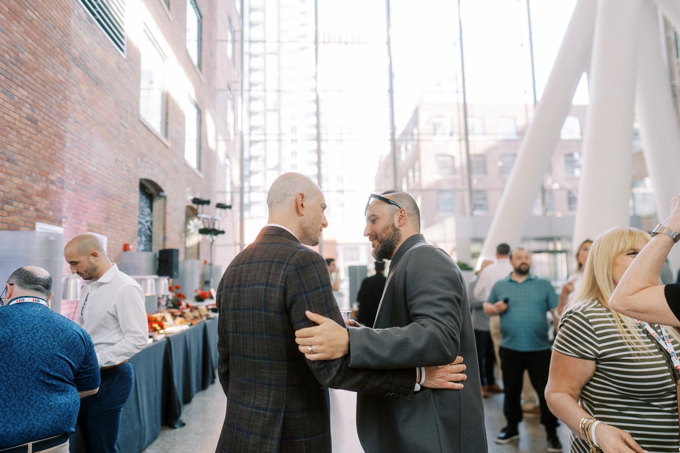 Two men greet each other and converse in the sunlit atrium of Ricarda’s during the Toronto Prep Forever Launch Party, with other guests mingling nearby.