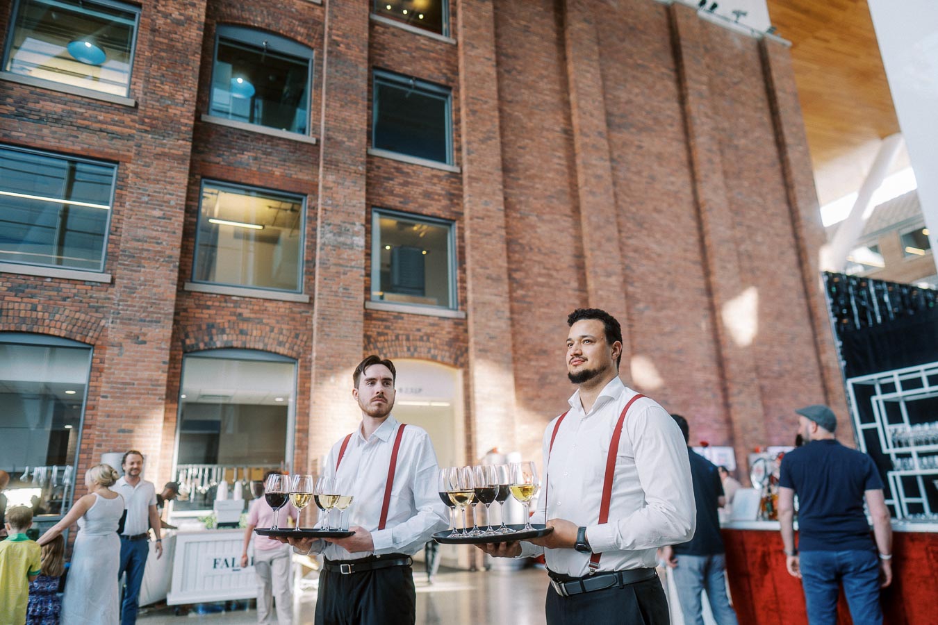 Two waitstaff in white shirts and red suspenders hold trays of wine glasses at Ricarda’s Atrium during the Toronto Prep Forever Launch Party, with guests and brick walls visible behind them.
