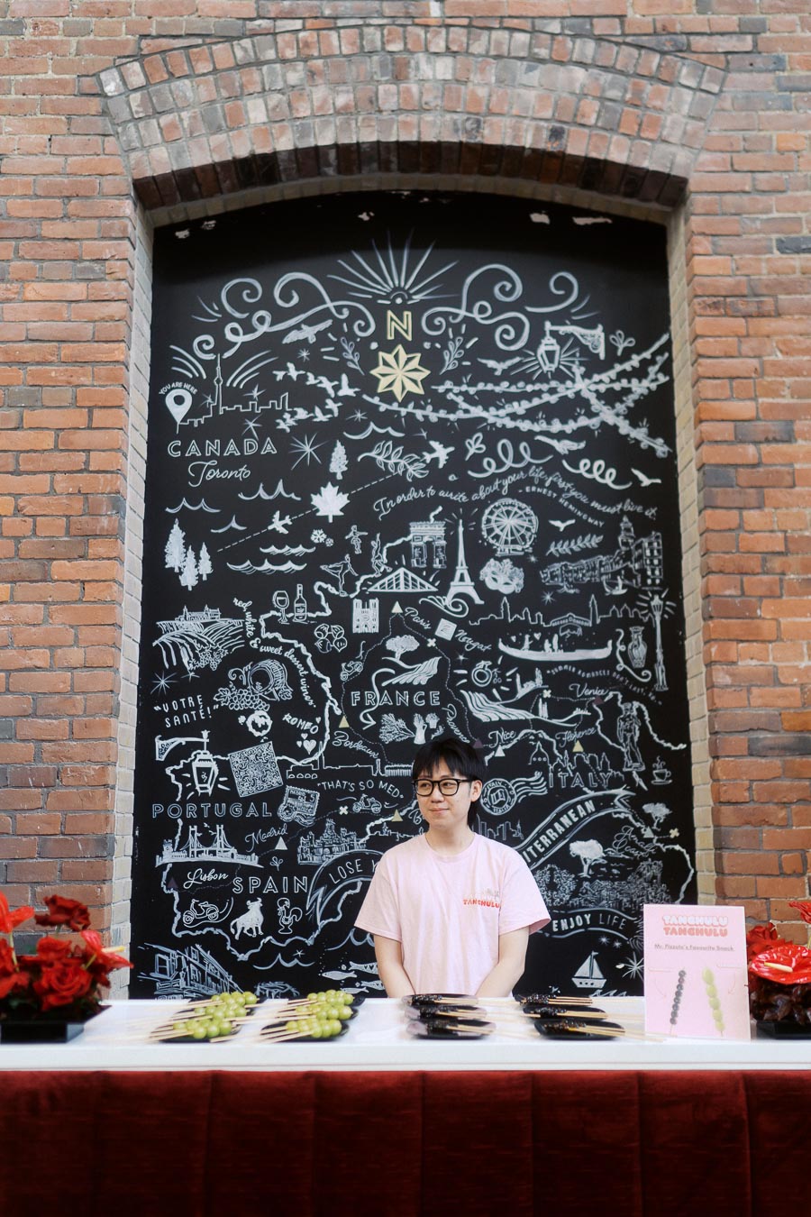 A Tanghulu vendor in a pink shirt stands behind a table of candied fruit skewers at Ricarda’s in Toronto, with a decorative chalk mural and brick wall in the background.