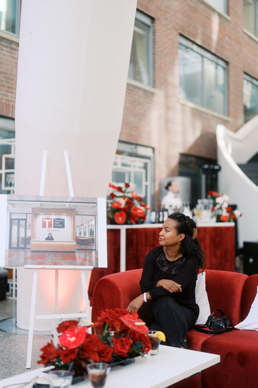 A woman sits on a red sofa near a rendering of Toronto Prep School at Ricarda’s Atrium, Toronto, with floral decor and event bar in the background.