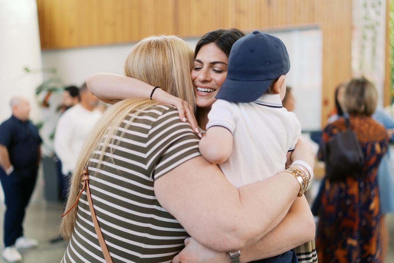 Two women warmly embrace while a young boy in a blue cap is held between them, surrounded by guests at Ricarda’s during the Toronto Prep Forever Launch Party.
