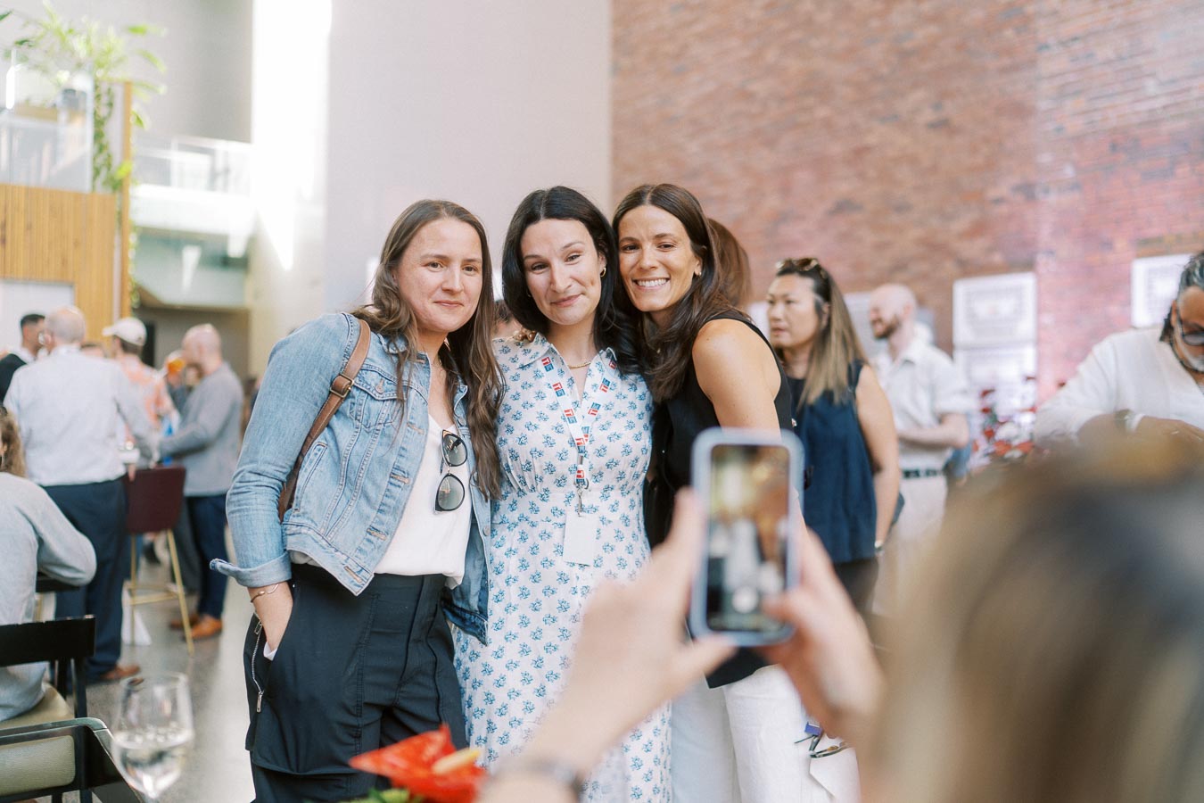 Three women stand close together and smile for a photo at the Toronto Prep Forever Launch Party at Ricarda’s, with guests socializing in the background.