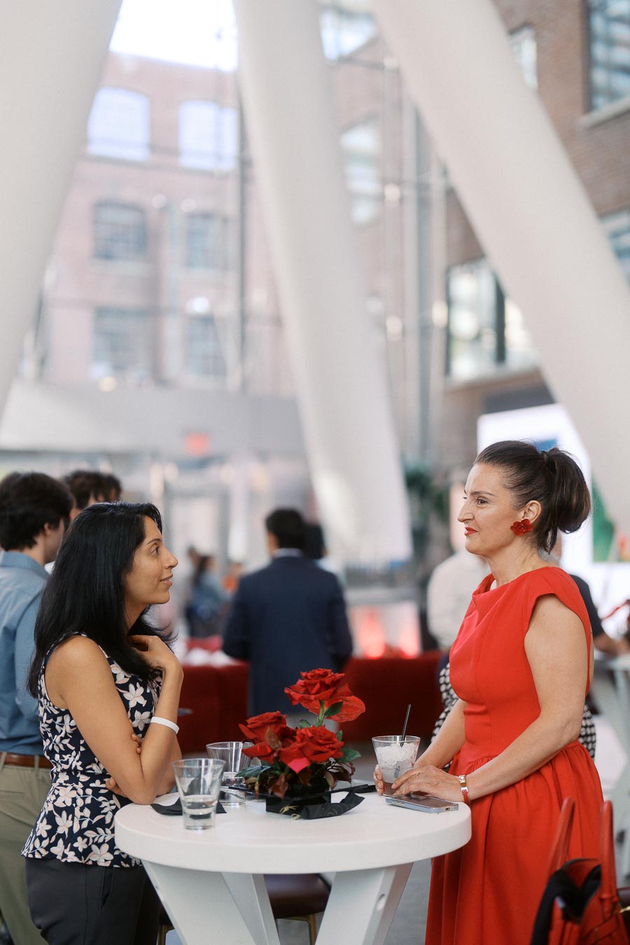 Two women talking at a round cocktail table with red flowers, inside The Atrium at Ricarda’s during the Toronto Prep Forever Launch Party.