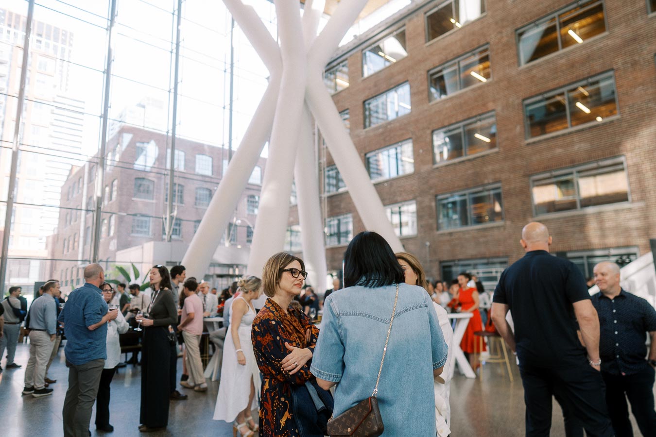 Guests gathering and socializing in the bright glass atrium of Ricarda’s, Toronto, during the Toronto Prep Forever Launch Party, with modern architecture and brick walls visible.