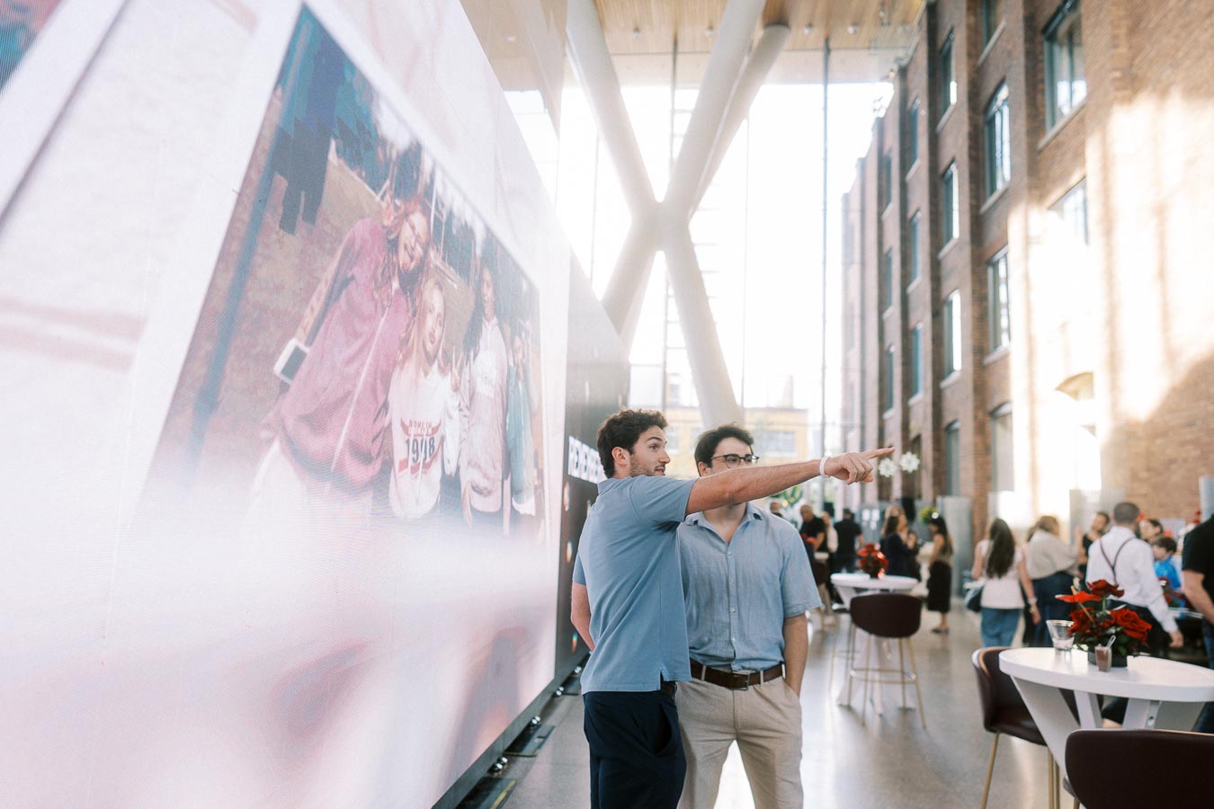 Two men stand in front of a large Toronto Prep School display, one pointing, in a bright atrium filled with guests at Ricarda’s in Toronto.