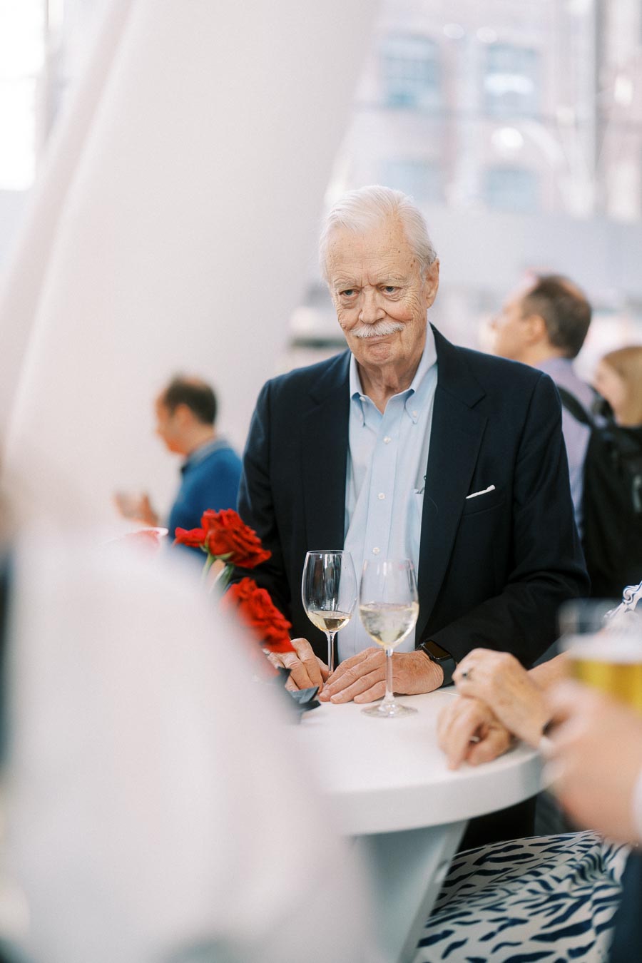 An older man in a suit stands at a white table with wine glasses and red flowers, mingling with other guests at the Toronto Prep Forever Launch Party at Ricarda’s in Toronto.