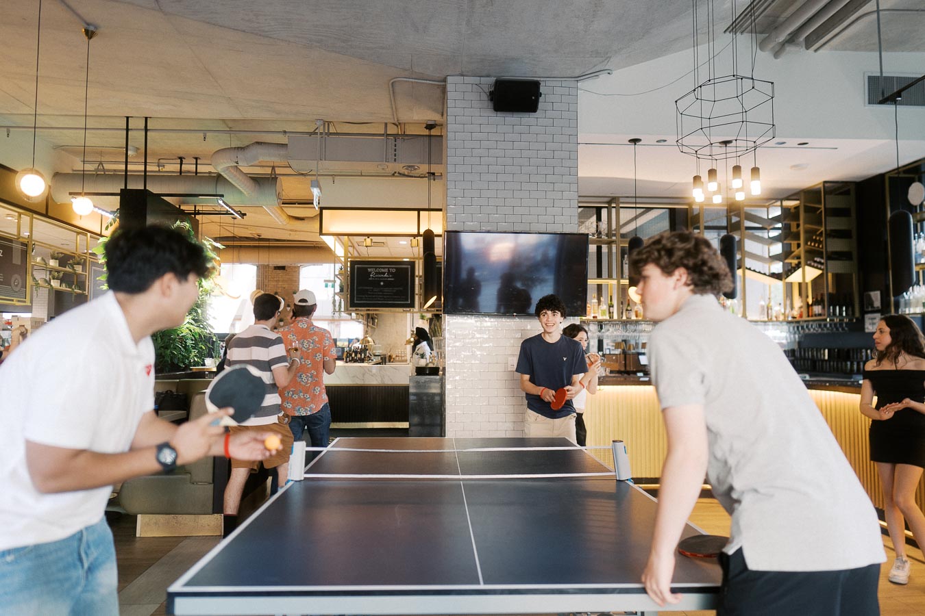 Two young people play ping pong at Ricarda’s in Toronto during the Toronto Prep Forever Launch Party, with others socializing in the background.