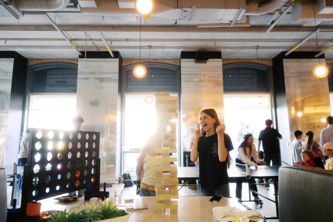A young girl celebrates during a giant Jenga game at the Toronto Prep Forever Launch Party at Ricarda’s, surrounded by other guests and games in a bright, modern event space.