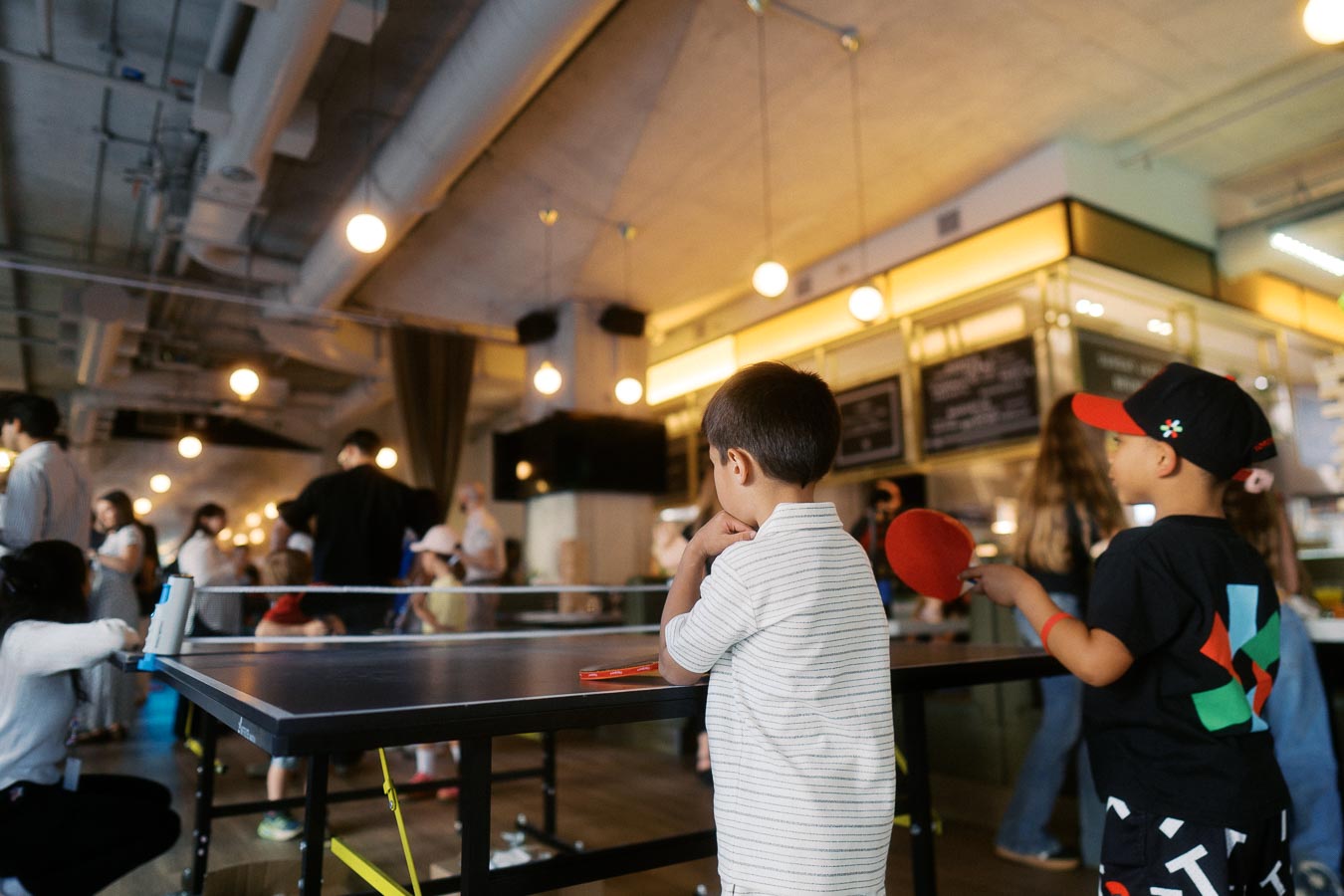 Two young boys play ping pong at a table in a lively indoor event space at Ricarda’s Toronto, with guests and families in the background.