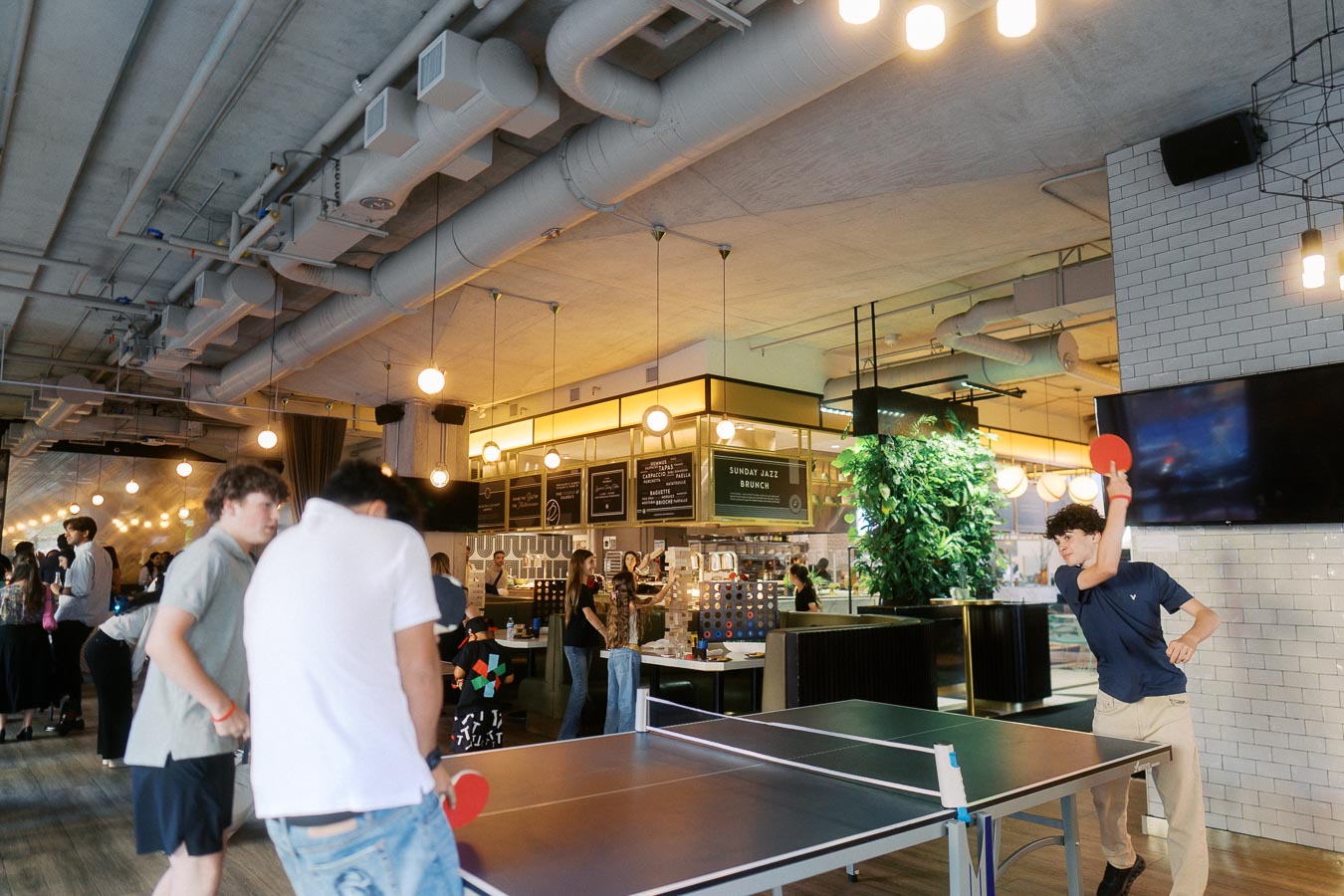 Three people play ping pong in a modern event space at Ricarda’s, Toronto, during the Toronto Prep School launch party, with guests mingling in the background.