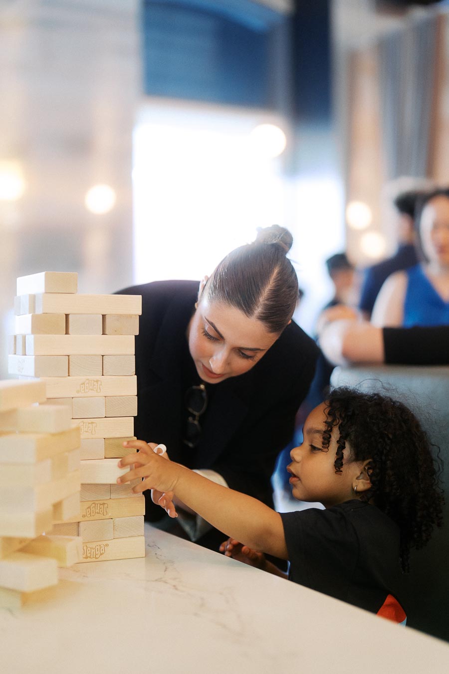 An adult and young child playing Jenga at a white table during the Toronto Prep Forever Launch Party at Ricarda’s, Toronto, with other guests in the background.