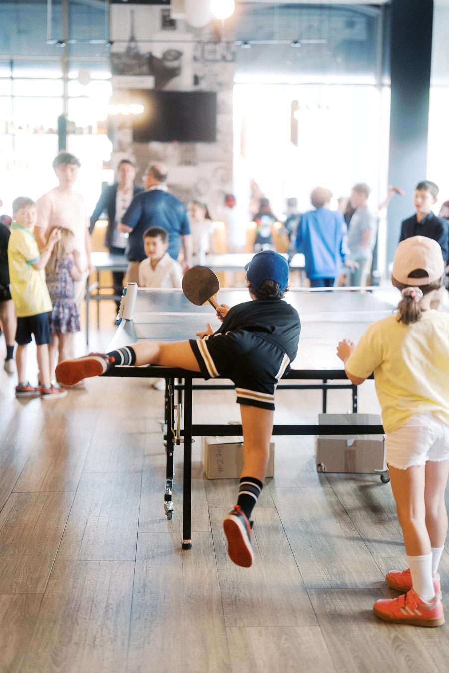 Two children play ping pong in a bright event space at Ricarda’s in Toronto, with other guests and families mingling in the background.