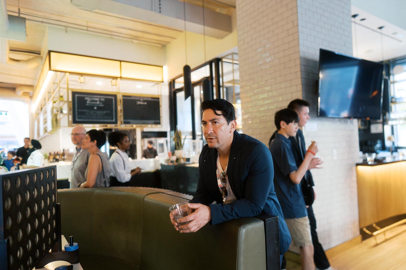 A man in a blue jacket leans on a booth holding a drink, while guests converse and staff work in the background at Ricarda’s, Toronto.