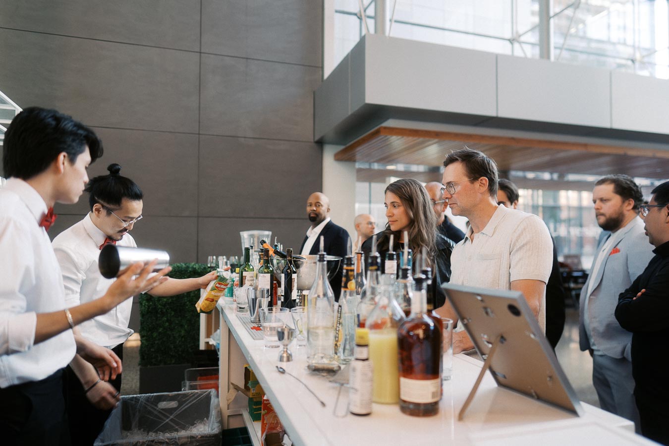 Guests interact with bartenders at the Ricarda’s bar during the Toronto Prep Forever Launch Party, with bottles and drink mixers on the counter in a modern atrium setting.