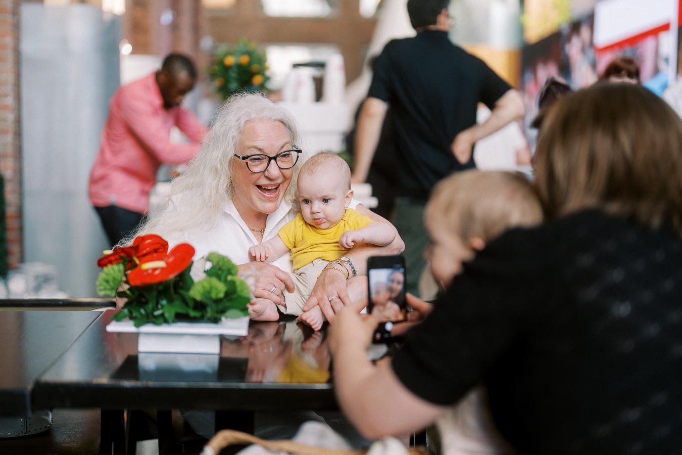 A smiling woman with long white hair and glasses holds a baby in a yellow shirt, while another adult photographs them at a table with flowers during a Toronto Prep School event at Ricarda’s in Toronto.