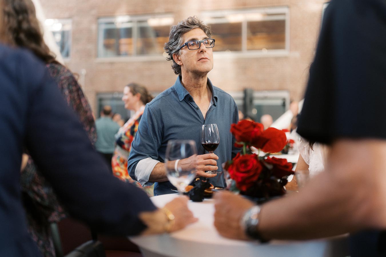 A man holding a glass of red wine at a cocktail table with red roses, mingling with other guests during the Toronto Prep School event at Ricarda’s Atrium.
