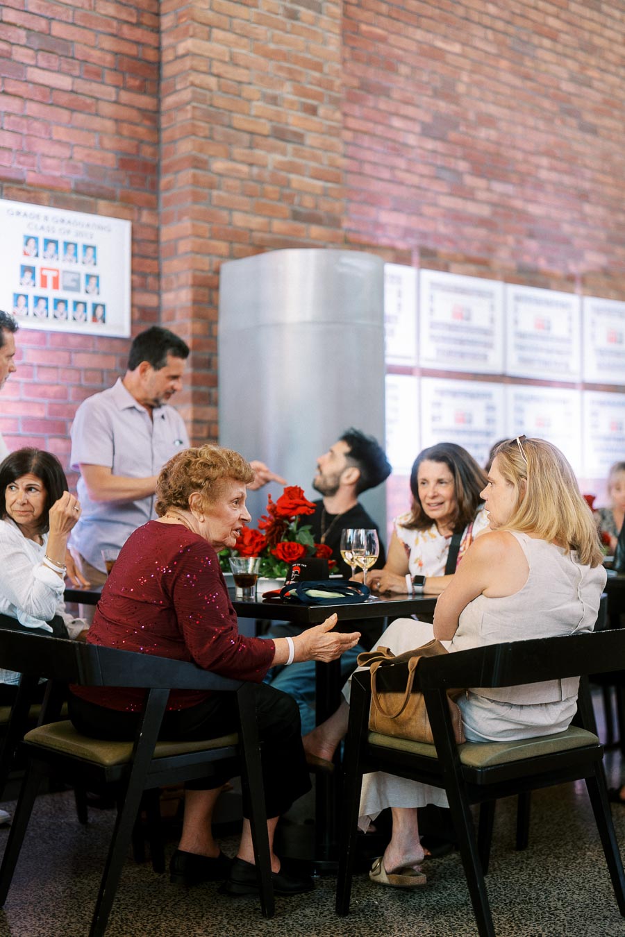 A group of guests seated around a table with drinks and red floral centrepieces, chatting in Ricarda’s Atrium at the Toronto Prep Forever Launch Party, with brick walls in the background.