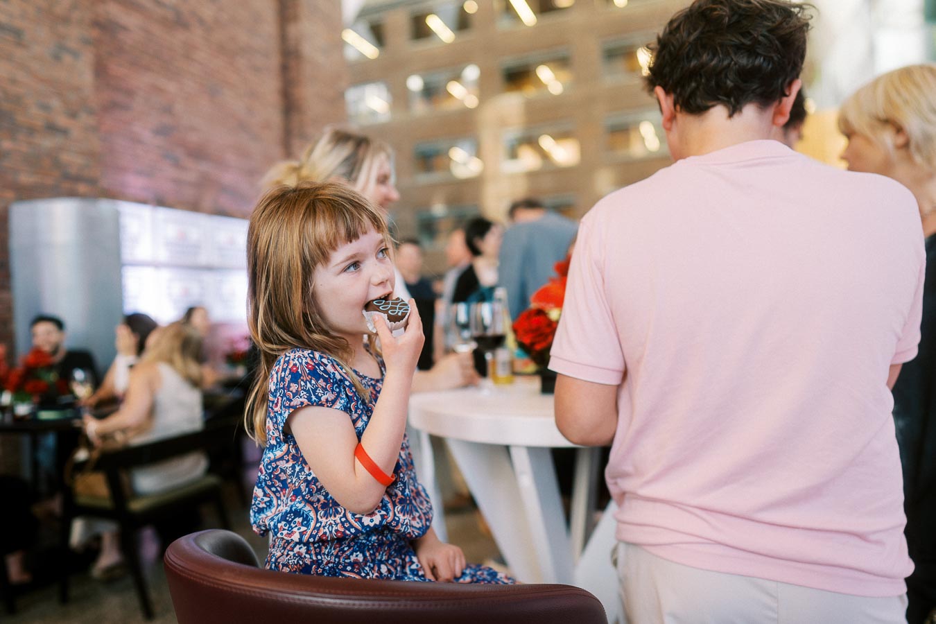 A young girl in a patterned dress eats a decorated cookie at a high-top table during the Toronto Prep Forever Launch Party at Ricarda’s in Toronto, with families and guests mingling in the background.