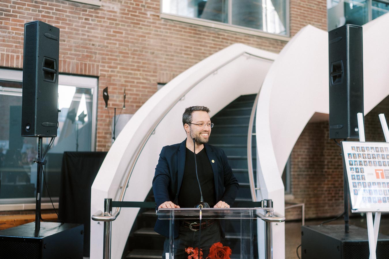 A man stands at a clear podium with a microphone, delivering a speech inside The Atrium at Ricarda’s in Toronto, with brick walls and a modern staircase in the background.