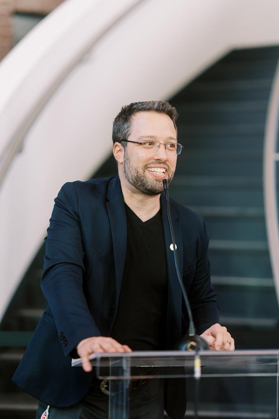 A man in a dark blazer speaks at a clear podium with a microphone, standing in front of a modern staircase at Ricarda’s in Toronto during the Toronto Prep Forever Launch Party.