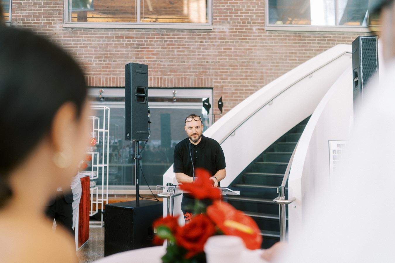 A man stands at a podium with a microphone in front of a curved staircase, addressing guests at the Toronto Prep Forever Launch Party at Ricarda’s in Toronto.