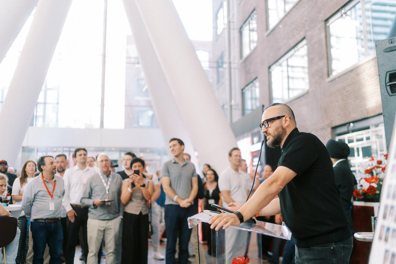 A man speaks at a podium to a gathered audience inside the modern glass atrium of Ricarda’s during the Toronto Prep School launch event.