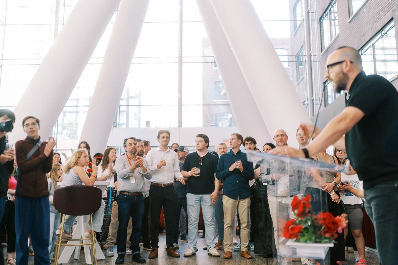 A diverse crowd listens to a speaker at a podium during the Toronto Prep Forever Launch Party at The Atrium at Ricarda’s, Toronto, with modern architecture in the background.