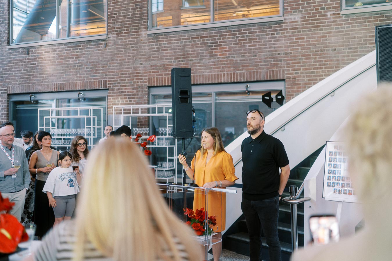 A woman speaks at a podium next to a man during the Toronto Prep Forever Launch Party at Ricarda’s in Toronto, with guests and families listening in a modern atrium setting.