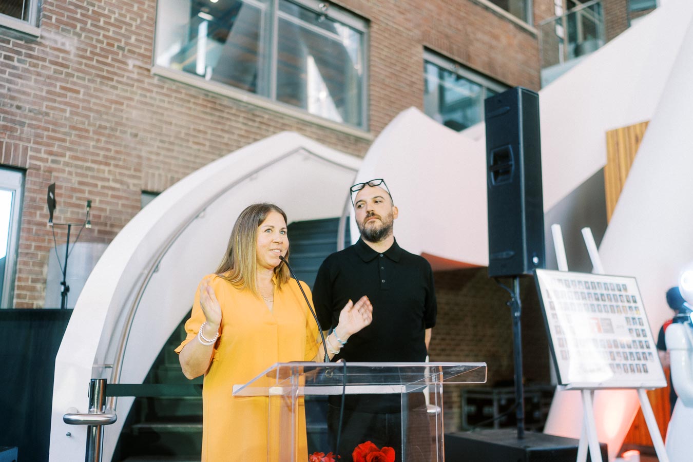 A woman in a yellow dress and a man in black stand behind a clear podium, speaking at the Toronto Prep Forever Launch Party at Ricarda’s, Toronto, with brick walls and event signage visible.