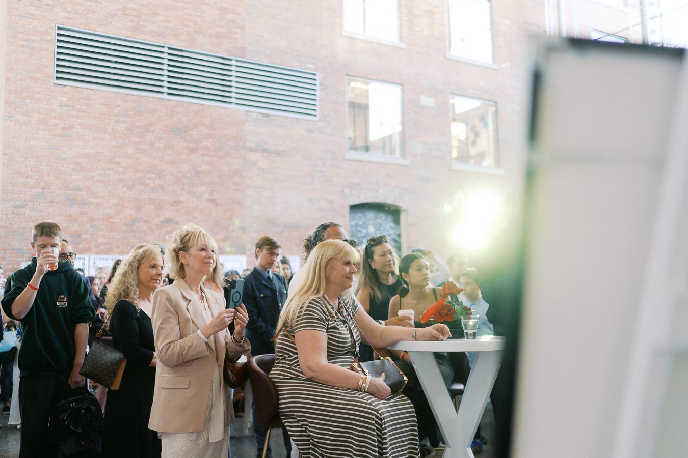 A group of guests, including women and teens, gathered around a cocktail table, attentively watching an event at The Atrium at Ricarda’s, Toronto Prep Forever Launch Party.